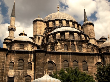 A historic mosque with multiple domes and tall minarets, constructed using stone and adorned with decorative elements. The architecture features intricate patterns on the window grilles and a blend of shadows and sunlight that highlights the texture of the stone. The sky is partly cloudy, adding depth to the scene.