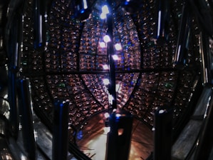 A circular metallic structure is filled with an array of small interconnected reflective surfaces. The structure is illuminated by bright lights, creating a play of light and shadows. The overall design resembles a complex technological or scientific apparatus, with shiny metal components and a symmetrical layout.