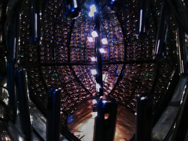 A circular metallic structure is filled with an array of small interconnected reflective surfaces. The structure is illuminated by bright lights, creating a play of light and shadows. The overall design resembles a complex technological or scientific apparatus, with shiny metal components and a symmetrical layout.