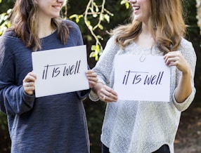 Two people are holding signs that read 'it is well' while smiling at each other. They are outdoors, with greenery visible in the background. The person on the left wears a dark blue sweater, and the one on the right has a light gray sweater.