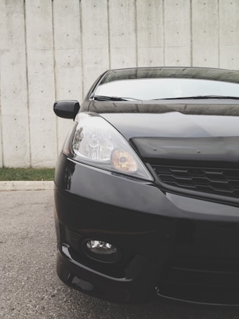 The front section of a sleek, black car is prominently displayed against a concrete wall backdrop. The image captures details of the car's headlight, grille, and hood with reflections visible on the polished surface.