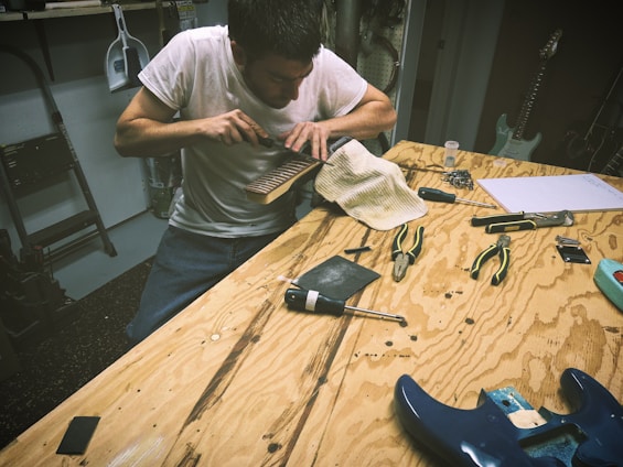 A person is working on a guitar neck using a tool on a wooden workbench. Various tools such as pliers, screwdrivers, and guitar components are scattered around. The setting appears to be a workshop or garage environment.