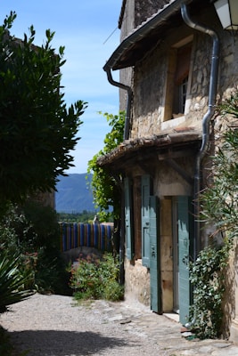 A narrow cobblestone alley lined with an old stone house featuring blue shutters. Lush greenery, including climbing vines and bushes, frames the scene. In the background, colorful striped fabric hangs over a railing, and distant mountains are visible under a clear blue sky.