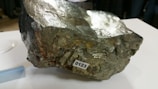 A large, rough metallic rock with reflective surfaces sits on a white table. It has a label with the number 0123 adhered to its surface, suggesting it is part of a collection or study. There are blurred figures in the background, indicating a possibly indoor setting, such as a museum or exhibition.