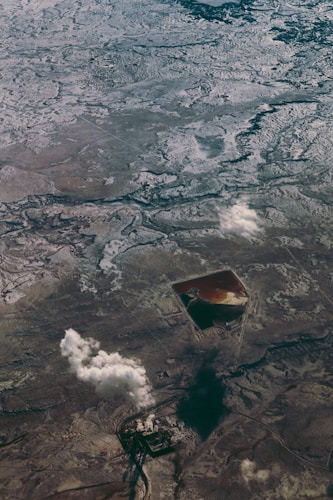 An aerial view of a vast, rugged terrain showing a combination of snow or ice-covered ground and brown earth. A geometrically shaped artificial structure, possibly a pond or mining pit, is visible, filled with liquid of different colors. There are scattered clouds casting shadows on the landscape below.