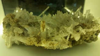 An intricate cluster of transparent, elongated crystal structures emerging from a rocky base, with various shades of beige and hints of green reflections. The crystals are densely packed and appear sharp and delicate.