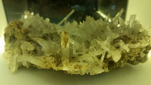 An intricate cluster of transparent, elongated crystal structures emerging from a rocky base, with various shades of beige and hints of green reflections. The crystals are densely packed and appear sharp and delicate.