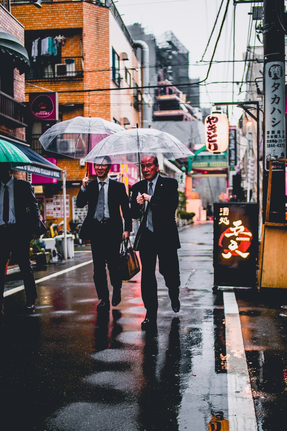 검은 양복을 입은 두 남자가 투명한 우산을 들고 거리를 걷고 있다