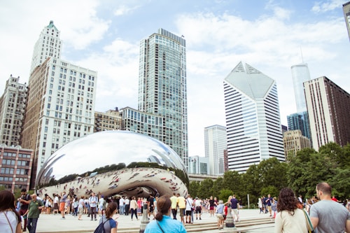 A large, reflective sculpture sits in a bustling plaza surrounded by modern skyscrapers. People of various ages are walking around and taking photos. The sculpture reflects the sky and diverse crowd, creating a dynamic scene. Lush green trees provide contrast against the urban backdrop.