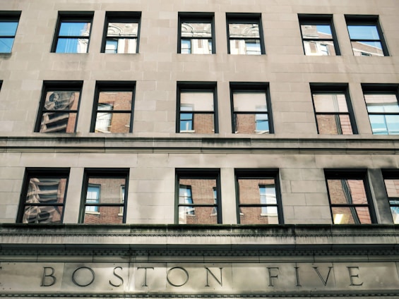 A stone facade of a building with multiple rectangular windows reflecting a different building with a similar architectural style. The name 'BOSTON FIVE' is engraved in large letters on the lower part of the facade. The reflections in the windows show parts of another structure with red brick walls and white window frames.