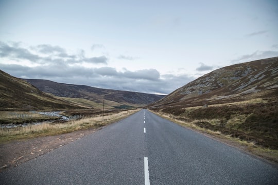 gray concrete road between green grass field near mountain under white clouds during daytime in Cairngorms National Park United Kingdom
