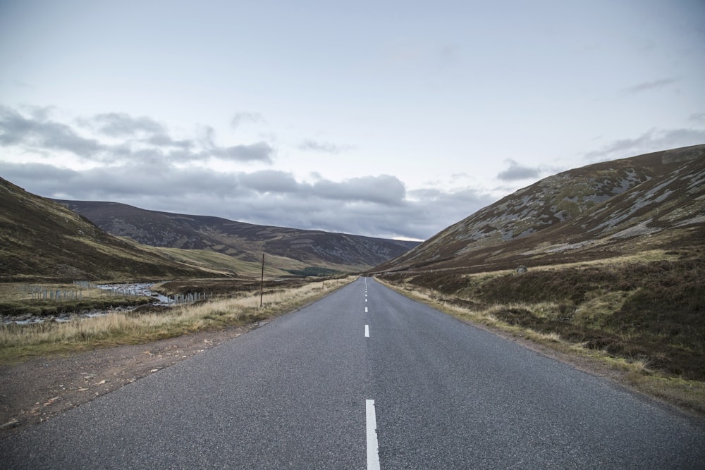 gray concrete road between green grass field near mountain under white clouds during daytime