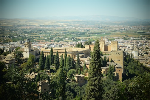 An expansive view of a historic cityscape with landmark architecture, including towers and fortress-like structures, surrounded by lush greenery and set against a backdrop of vast plains and distant mountains.