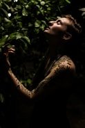 A person with leaf-patterned tattoos on their arm stands among lush green foliage. The lighting is low and dramatic, highlighting their serene expression as they tilt their head back, possibly enjoying the natural surroundings.