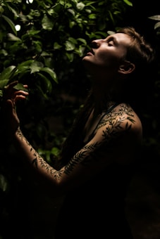 A person with leaf-patterned tattoos on their arm stands among lush green foliage. The lighting is low and dramatic, highlighting their serene expression as they tilt their head back, possibly enjoying the natural surroundings.