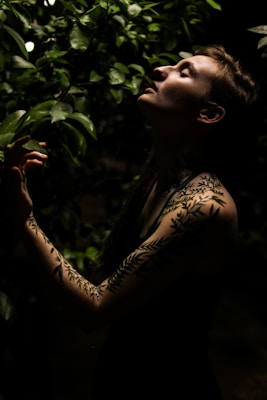 A person with leaf-patterned tattoos on their arm stands among lush green foliage. The lighting is low and dramatic, highlighting their serene expression as they tilt their head back, possibly enjoying the natural surroundings.
