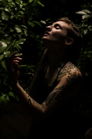 A person with intricate leaf tattoos stands in a shadowy, verdant setting, reaching toward the light filtering through the leaves. The scene is serene and intimate, with a focus on the interplay of light and shadow on the person's face and body.