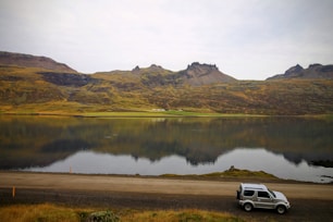A serene landscape featuring a calm lake reflecting rugged, green hills and distant mountains. A small SUV is parked on the roadside in the foreground, adding a sense of adventure and exploration to the natural scene.