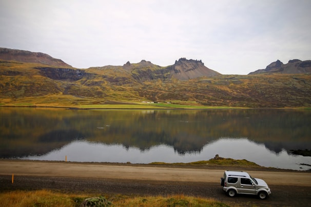 A serene landscape featuring a calm lake reflecting rugged, green hills and distant mountains. A small SUV is parked on the roadside in the foreground, adding a sense of adventure and exploration to the natural scene.