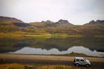 A serene landscape featuring a calm lake reflecting rugged, green hills and distant mountains. A small SUV is parked on the roadside in the foreground, adding a sense of adventure and exploration to the natural scene.