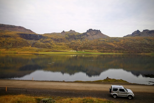 A serene landscape featuring a calm lake reflecting rugged, green hills and distant mountains. A small SUV is parked on the roadside in the foreground, adding a sense of adventure and exploration to the natural scene.
