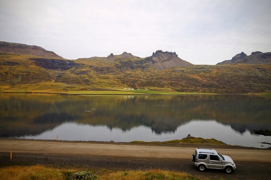 A serene landscape featuring a calm lake reflecting rugged, green hills and distant mountains. A small SUV is parked on the roadside in the foreground, adding a sense of adventure and exploration to the natural scene.