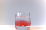 A clear glass filled with water. A red substance appears to be partially mixed in the water, creating a gradient effect from red to clear. The background is a soft gradient of light grey to white, and the glass is placed on a white surface.