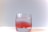 A clear glass filled with water. A red substance appears to be partially mixed in the water, creating a gradient effect from red to clear. The background is a soft gradient of light grey to white, and the glass is placed on a white surface.