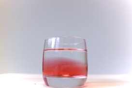 A clear glass filled with water. A red substance appears to be partially mixed in the water, creating a gradient effect from red to clear. The background is a soft gradient of light grey to white, and the glass is placed on a white surface.