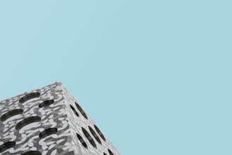 A modern building facade features a geometric pattern with arches and a mosaic-like design. The structure seems to be part of a larger building set against a clear blue sky, giving a minimalist aesthetic.