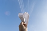 A hand holding a lined paper airplane against a clear blue sky with some faint cloud trails visible. The sunlight is casting a natural glow on the paper.