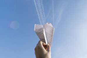 A hand holding a lined paper airplane against a clear blue sky with some faint cloud trails visible. The sunlight is casting a natural glow on the paper.