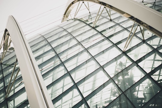 A close-up photo featuring a modern architectural structure characterized by large, curved glass panels. The structure includes a framework of metal beams and arches, showcasing an intricate and futuristic design.
