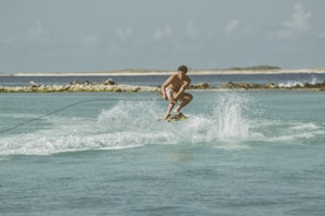 A person engaged in wakeboarding over a calm blue-green sea with waves and water splashes, holding onto a rope. The backdrop features a coastline with some distant hills and a clear sky.