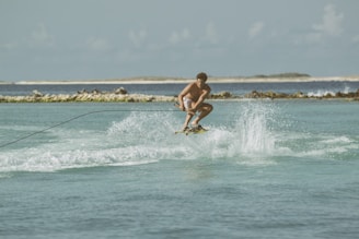 A person engaged in wakeboarding over a calm blue-green sea with waves and water splashes, holding onto a rope. The backdrop features a coastline with some distant hills and a clear sky.