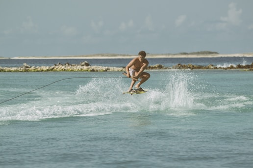 A person engaged in wakeboarding over a calm blue-green sea with waves and water splashes, holding onto a rope. The backdrop features a coastline with some distant hills and a clear sky.