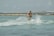A person engaged in wakeboarding over a calm blue-green sea with waves and water splashes, holding onto a rope. The backdrop features a coastline with some distant hills and a clear sky.