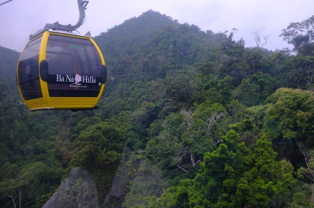 A yellow cable car hangs against a backdrop of lush green forest and misty mountain scenery, branded with 'Ba Na Hills' and 'Mountain resort' on its side.
