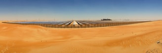Vast solar panel array stretches across a desert landscape, with sand dunes in the foreground and a clear blue sky overhead. The symmetry and alignment of the panels contrast with the organic forms of the surrounding sand.