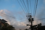 Utility poles with transformers and numerous power lines extending in various directions against a backdrop of a partly cloudy sky. The lower portion features sections of buildings and tree foliage.