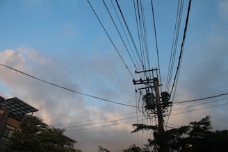 Utility poles with transformers and numerous power lines extending in various directions against a backdrop of a partly cloudy sky. The lower portion features sections of buildings and tree foliage.