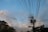 Utility poles with transformers and numerous power lines extending in various directions against a backdrop of a partly cloudy sky. The lower portion features sections of buildings and tree foliage.
