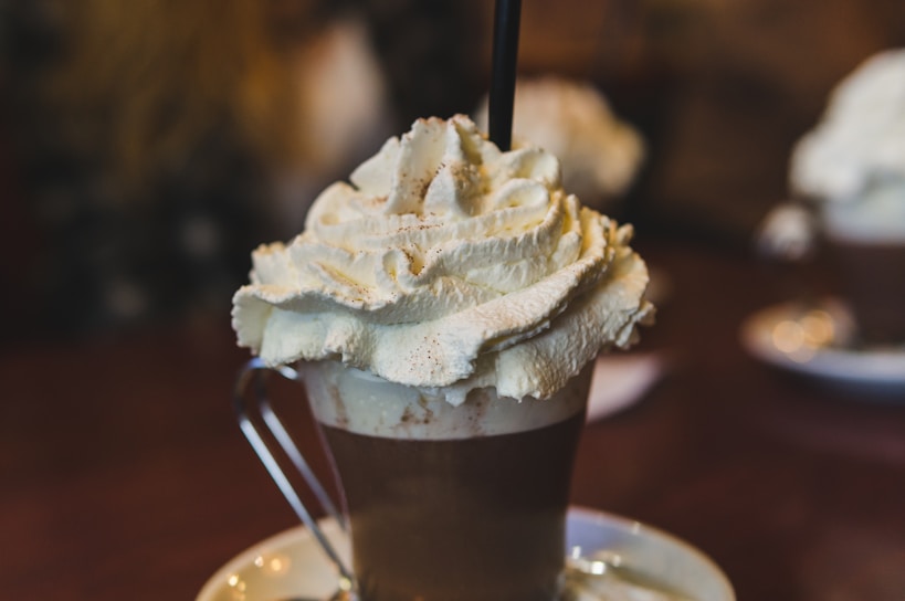 A glass mug filled with a hot beverage, topped with a generous amount of whipped cream and a straw. The background features an out-of-focus setting, hinting at a cozy indoor environment possibly in a cafe.