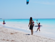 People are engaged in kitesurfing and walking along a sandy beach with clear turquoise water under a bright blue sky. A bridge is visible in the distance.