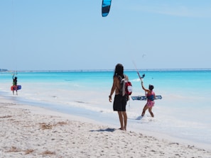 People are engaged in kitesurfing and walking along a sandy beach with clear turquoise water under a bright blue sky. A bridge is visible in the distance.