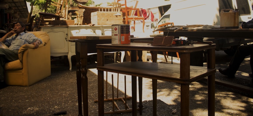 A person is sitting on a yellow armchair at an outdoor workshop area. Various items, including wooden furniture, tools, and paint cans, are scattered around. There is a van in the background, and the setting suggests a work or restoration area.