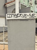 A concrete wall with a textured surface is prominently featured, with the words 'CONSERVE-SE' painted in bold, black lettering. In the background, the facade of a residential building is visible, painted in pastel green and gray tones, accented by red roof tiles. A metal fence is partially visible, alongside a paved walkway.