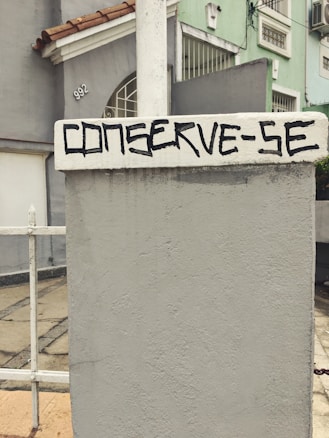 A concrete wall with a textured surface is prominently featured, with the words 'CONSERVE-SE' painted in bold, black lettering. In the background, the facade of a residential building is visible, painted in pastel green and gray tones, accented by red roof tiles. A metal fence is partially visible, alongside a paved walkway.