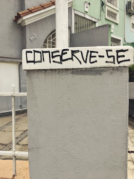 A concrete wall with a textured surface is prominently featured, with the words 'CONSERVE-SE' painted in bold, black lettering. In the background, the facade of a residential building is visible, painted in pastel green and gray tones, accented by red roof tiles. A metal fence is partially visible, alongside a paved walkway.