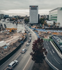 An urban construction site with several vehicles on a road running alongside. There are cranes and construction equipment visible at work within the enclosed site, surrounded by fencing. Tall buildings are present in the background with cloudy skies above.